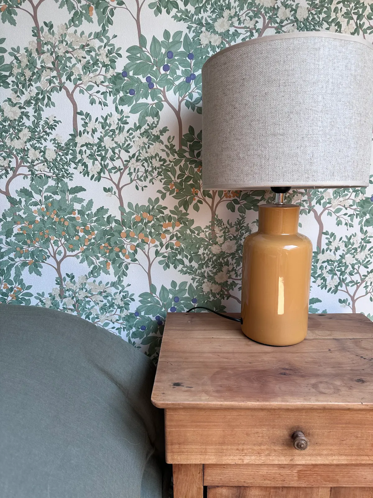 Papier peint représentant des arbres orangers derrière la tête de lit, une table de nuit ancienne et une lampe